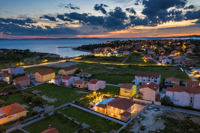 Osvijetljena dovršena vila s bazenom snimljena iz zraka u sumrak, iza nje naselje, uvala i more