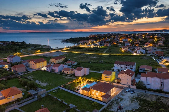 Osvijetljena dovršena vila s bazenom snimljena iz zraka u sumrak, iza nje naselje, uvala i more